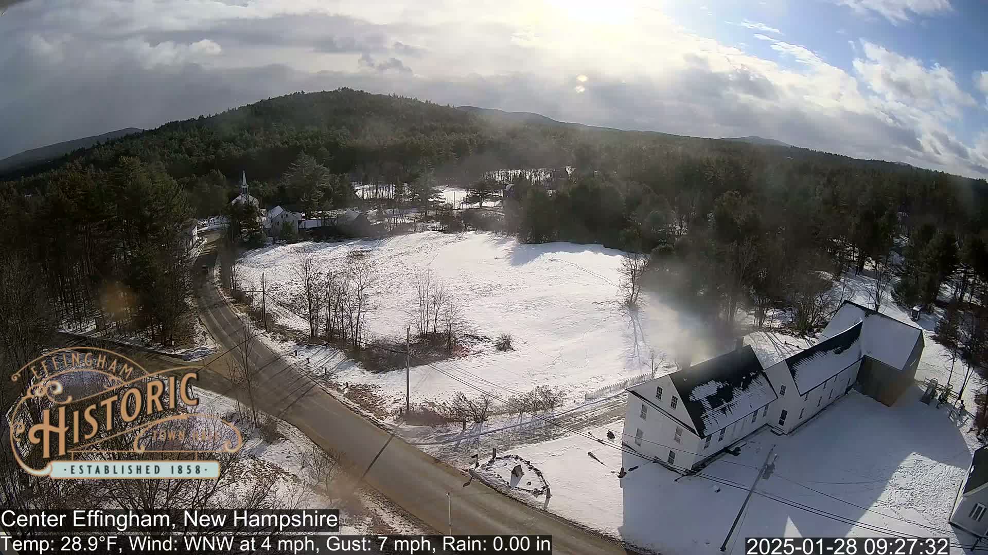 A snow-covered town, viewed from above on a partly sunny day with a few clouds, shows a road intersecting a field and several buildings, including a church.