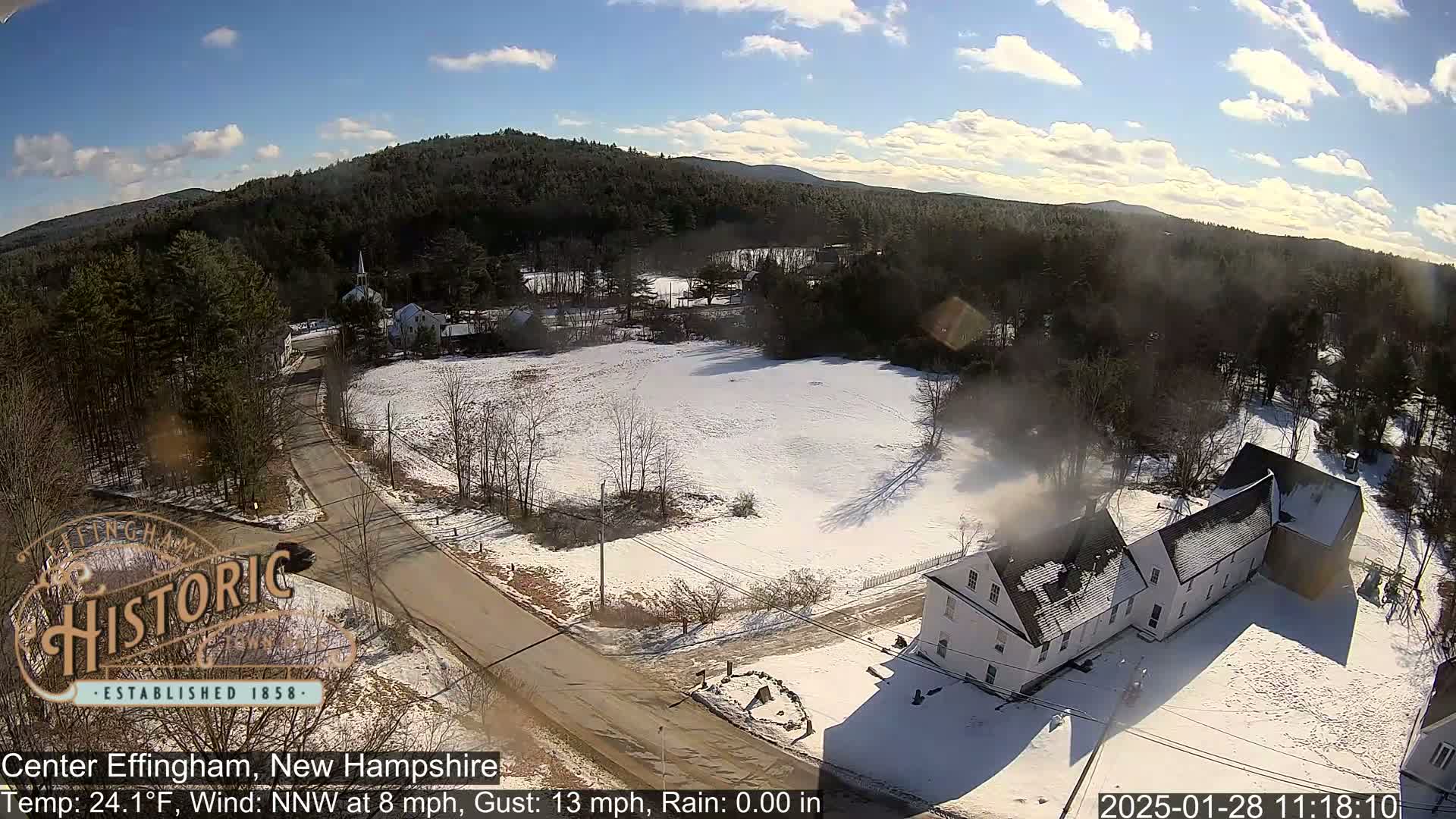 A partly sunny, snowy, rural landscape shows a road, buildings, and trees under a mostly clear sky.