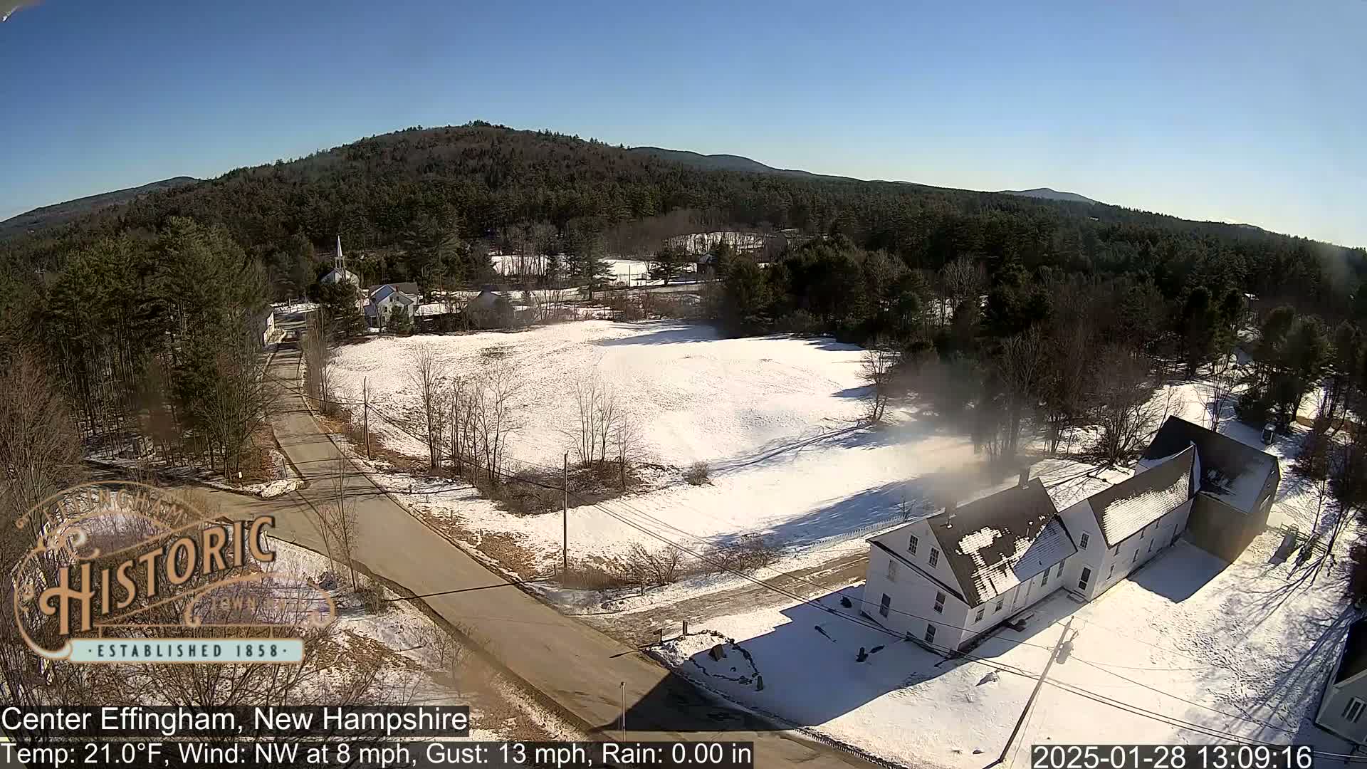 A snow-covered village scene with a road, buildings, and forested hills under a clear, sunny sky.