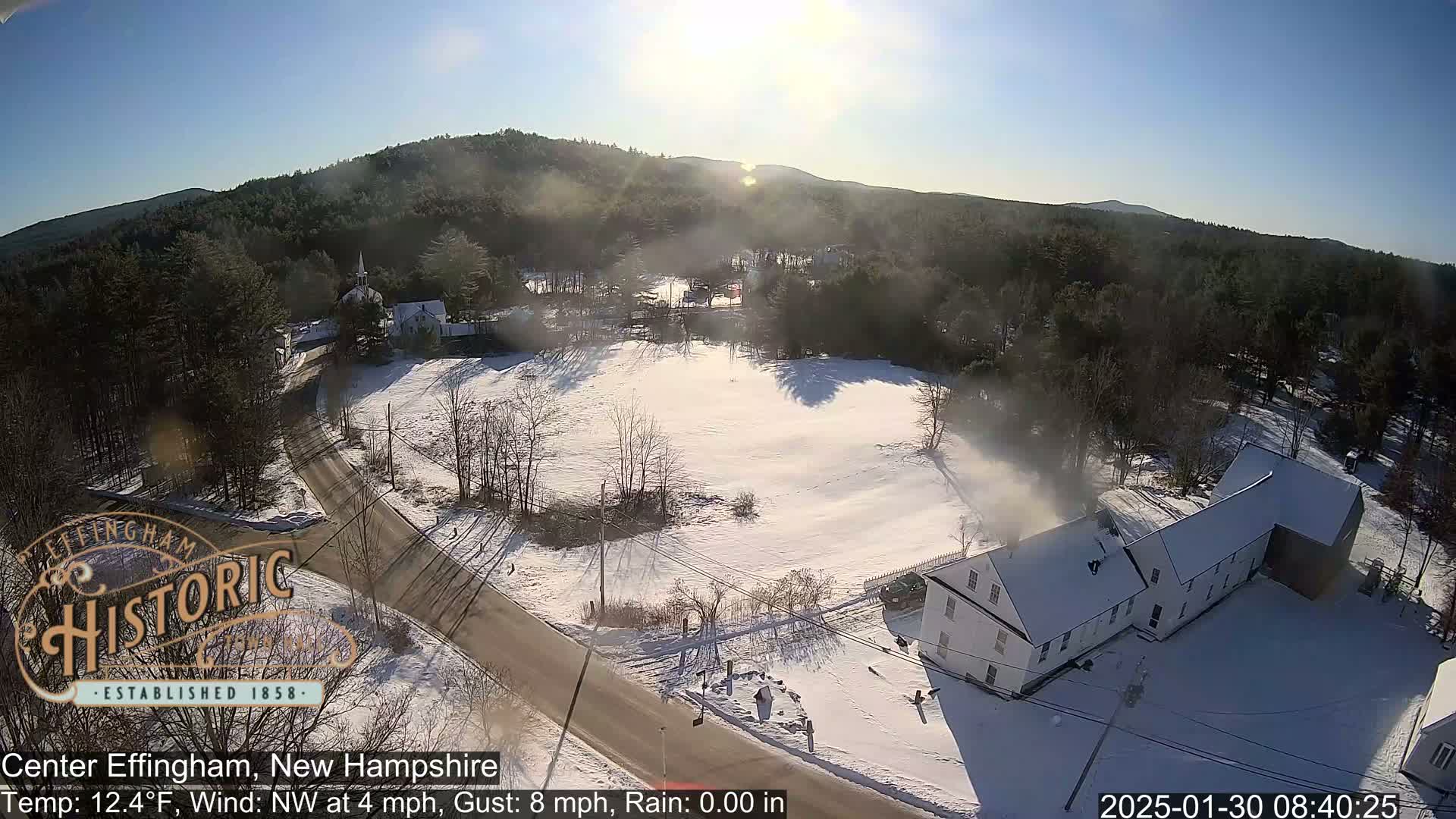 A snow-covered town, nestled among wooded hills, is viewed from above on a sunny, cold day.