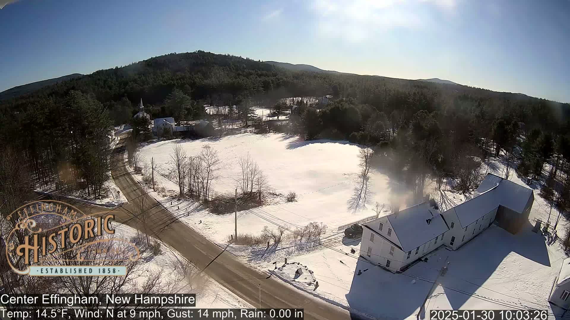 A snow-covered rural landscape with a road, buildings, and trees under a sunny sky.