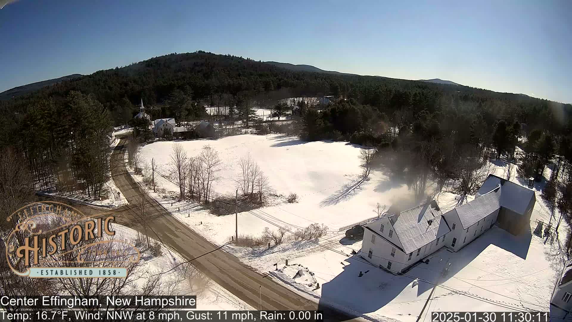 A snow-covered road winds through a small town nestled among snow-covered fields and evergreen trees under a bright, sunny sky.