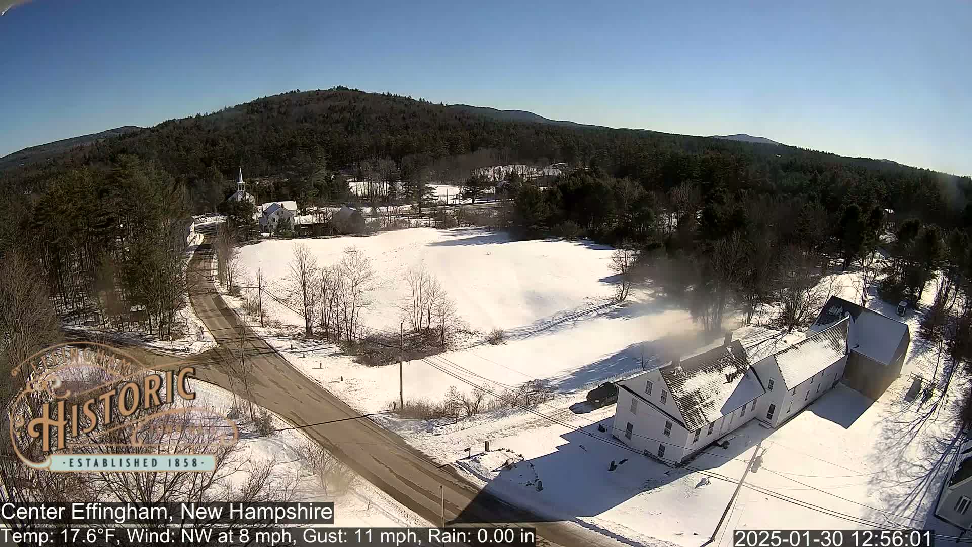 A snow-covered rural landscape with a road, buildings, and a church steeple, under a clear blue sky.