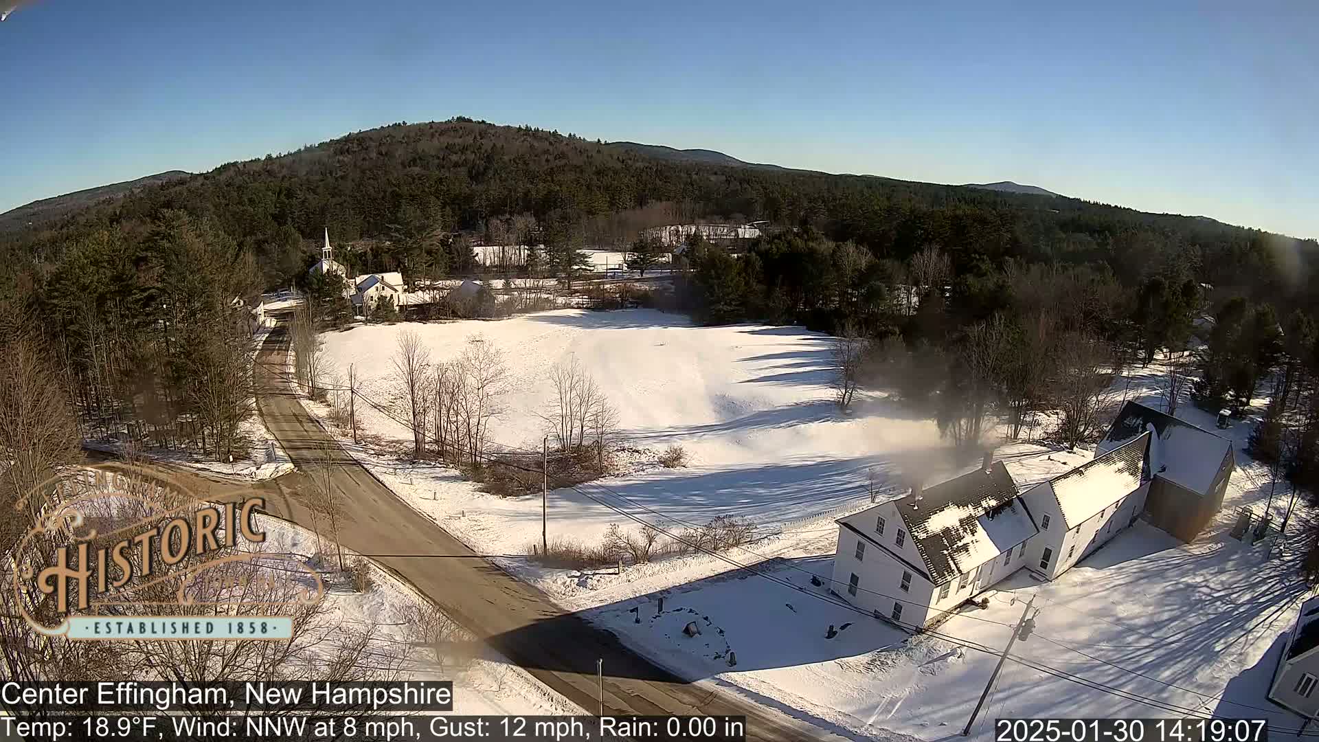 A snow-covered road leads to a small village nestled in a valley, with a church steeple visible in the distance, under a clear, sunny sky.