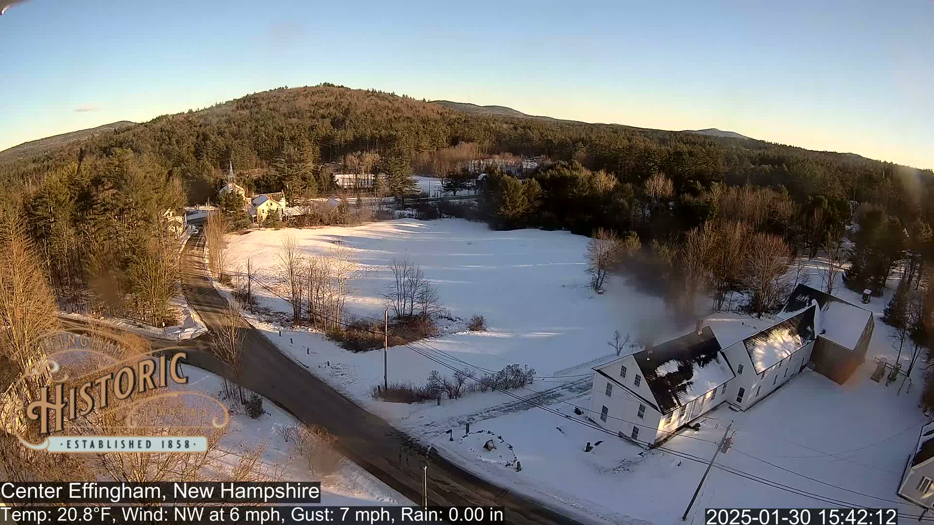 A snow-covered landscape with a village nestled among bare trees and a hill in the background under a clear, sunny sky.