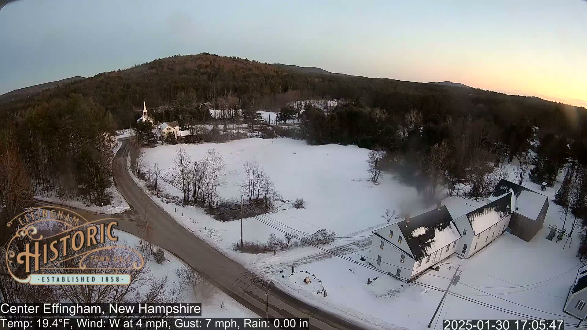 An aerial view shows a snow-covered town with houses and a church, under a twilight sky with a temperature of 19.4°F, a westerly wind at 4 mph, and gusts up to 7 mph.