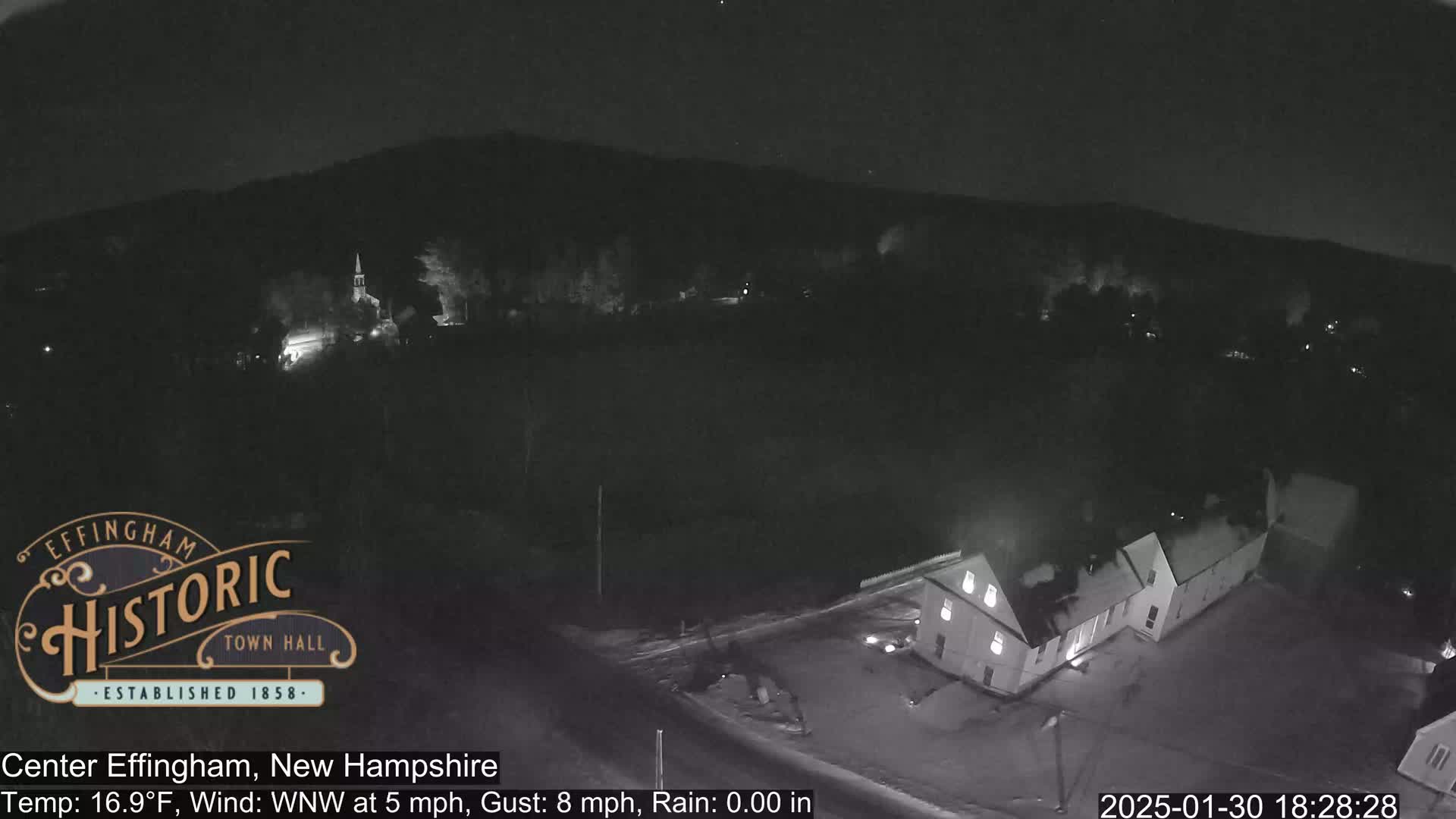 A nighttime, snowy scene shows a small town with a few lit buildings, including a long, low building near the bottom and a church-like structure further away in the background, under a dark, clear sky.