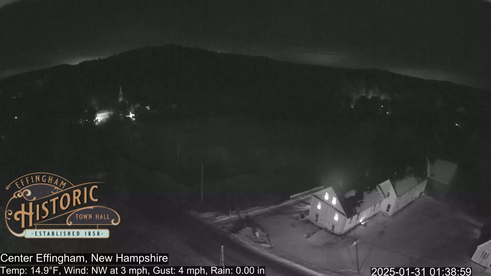A nighttime, low-light aerial view of a small town with a few buildings visible under a dark sky, with a slight northwest wind.