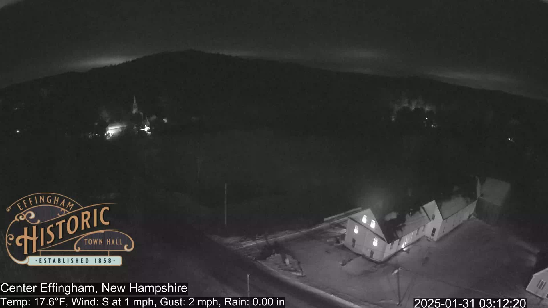 A nighttime, aerial view of a small town under rainy conditions shows a few buildings illuminated, with a dark, hilly landscape in the background.