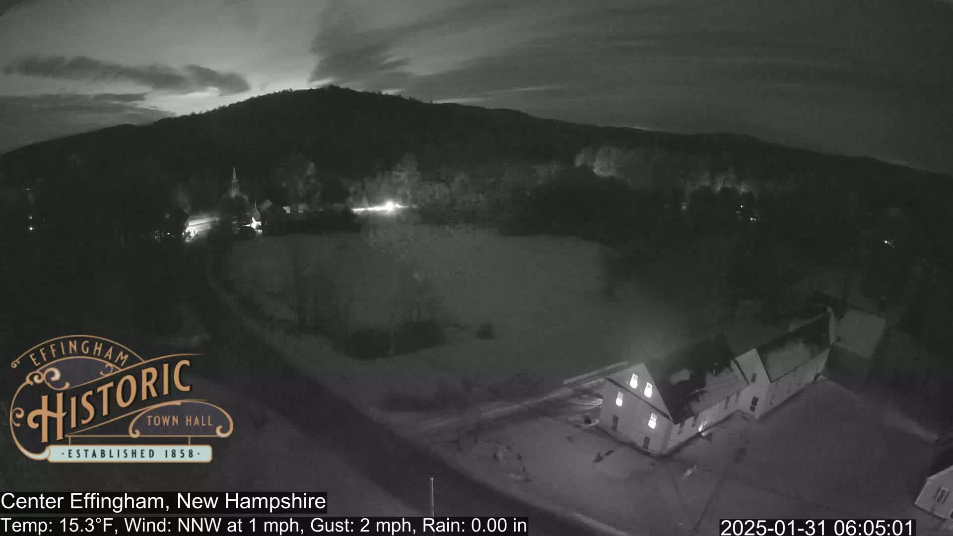 A nighttime, low-light view of a small town nestled against a dark hill under a mostly clear sky, with a light wind and no rain.
