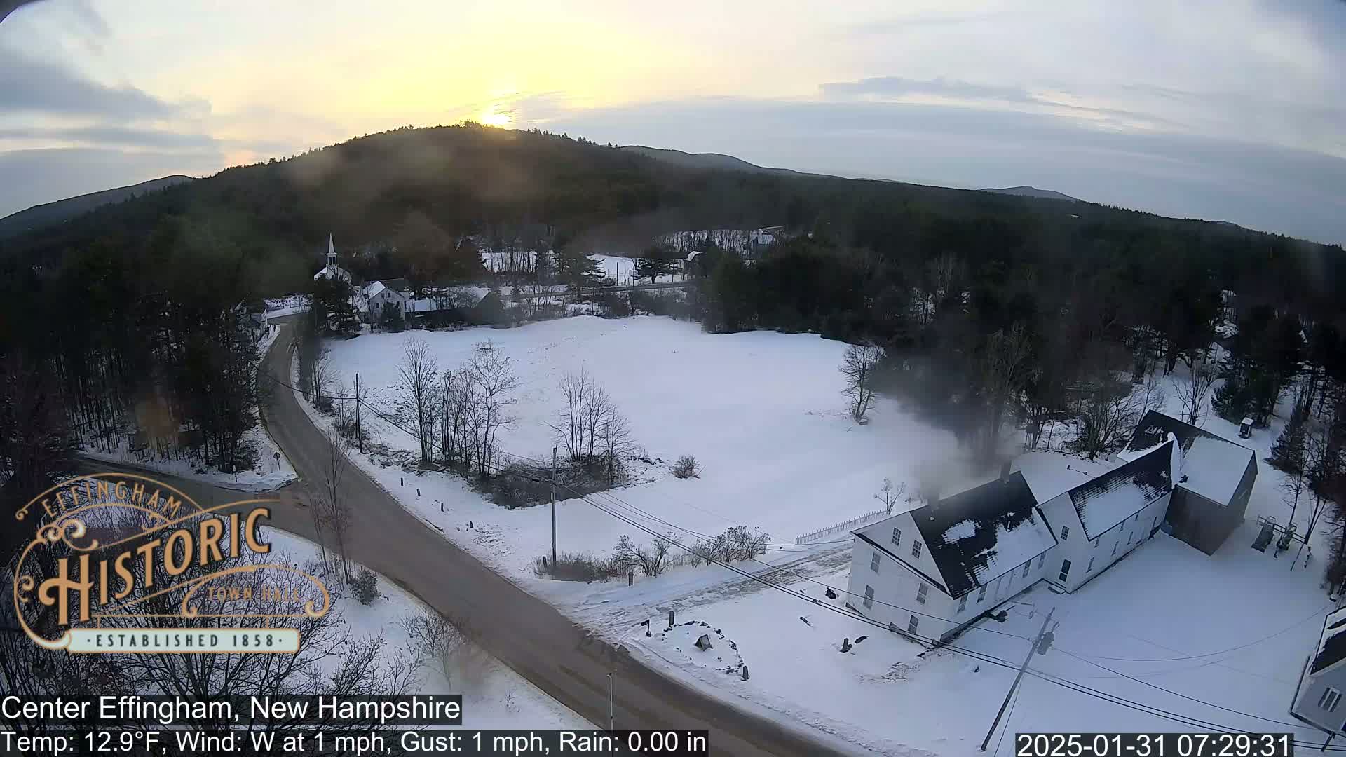 A snow-covered town at sunrise, with a road winding through the landscape and buildings visible among the trees.