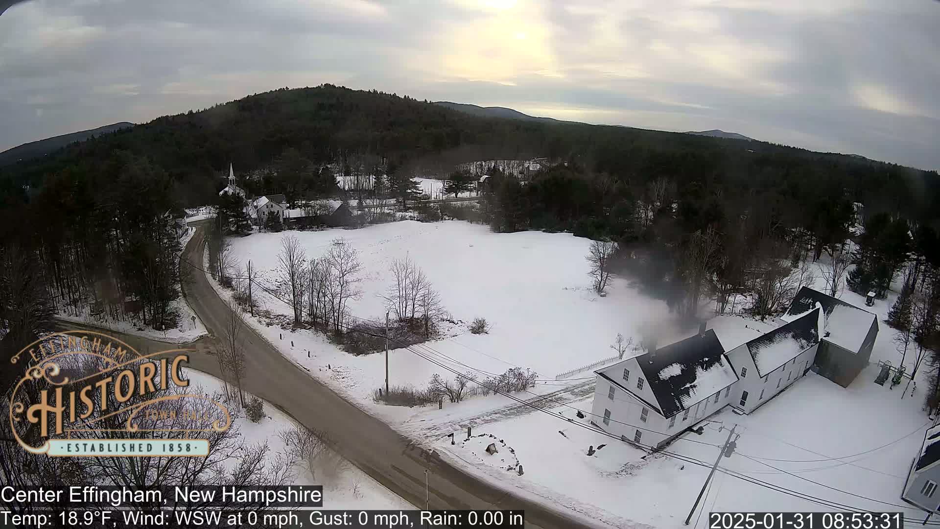 A snow-covered rural landscape with a road, buildings, and trees under an overcast sky.