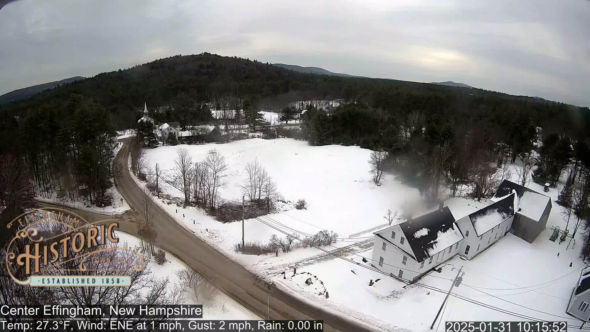 A snow-covered village scene with a few buildings and a road, viewed from above in overcast conditions.