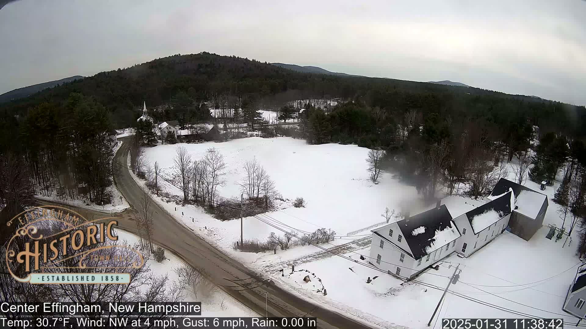 A snow-covered rural area with a road and several buildings is viewed from above on an overcast day.