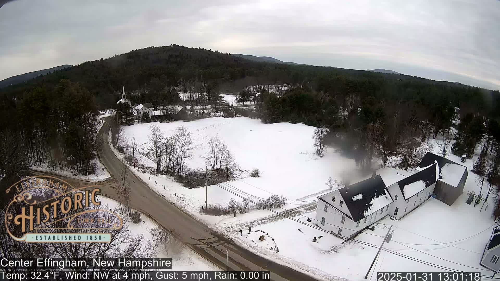 A snow-covered town in a wooded area, viewed from above on an overcast day with a light northwest wind.