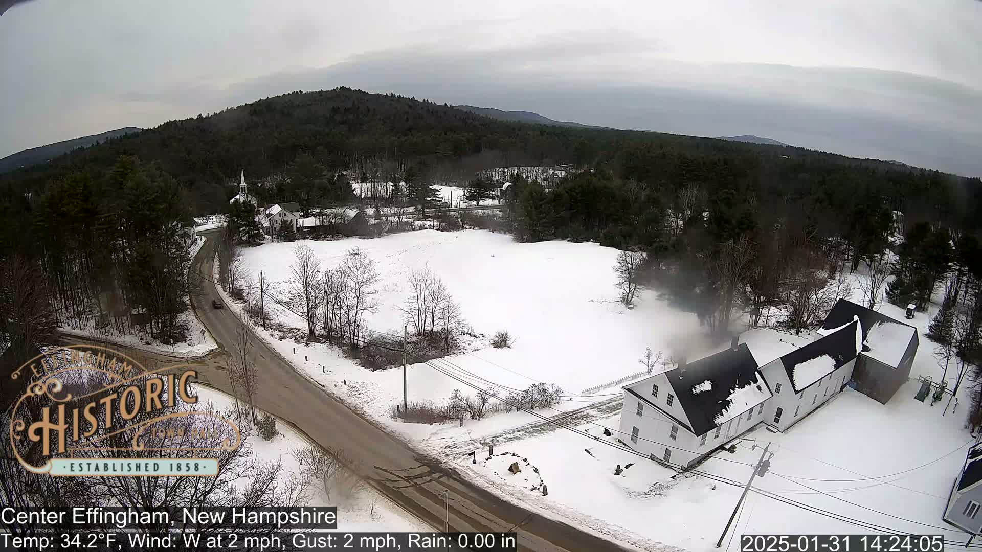 A snow-covered town in a rural area with a few buildings, a road, and a background of dark, wooded hills under an overcast sky.