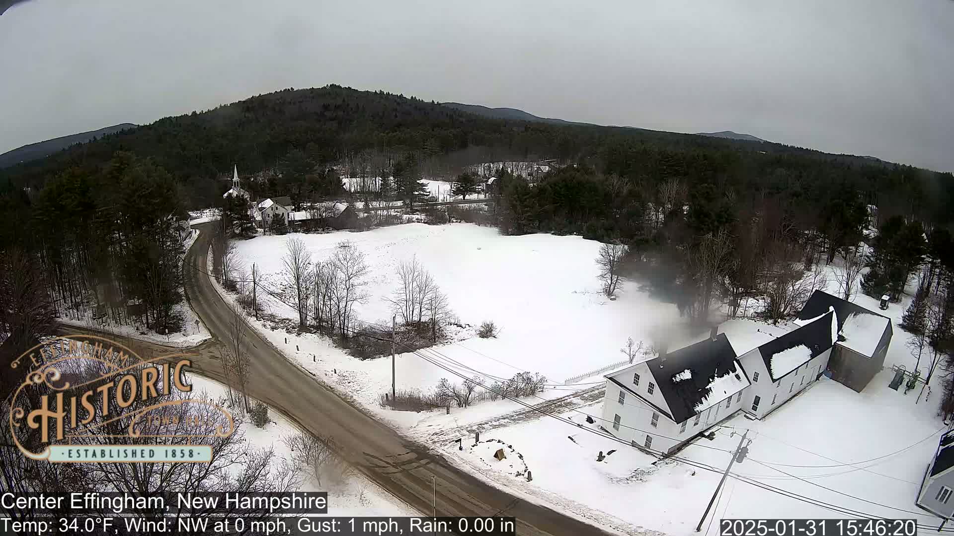 A snow-covered town scene with a road intersection and several buildings is viewed from above on an overcast day.