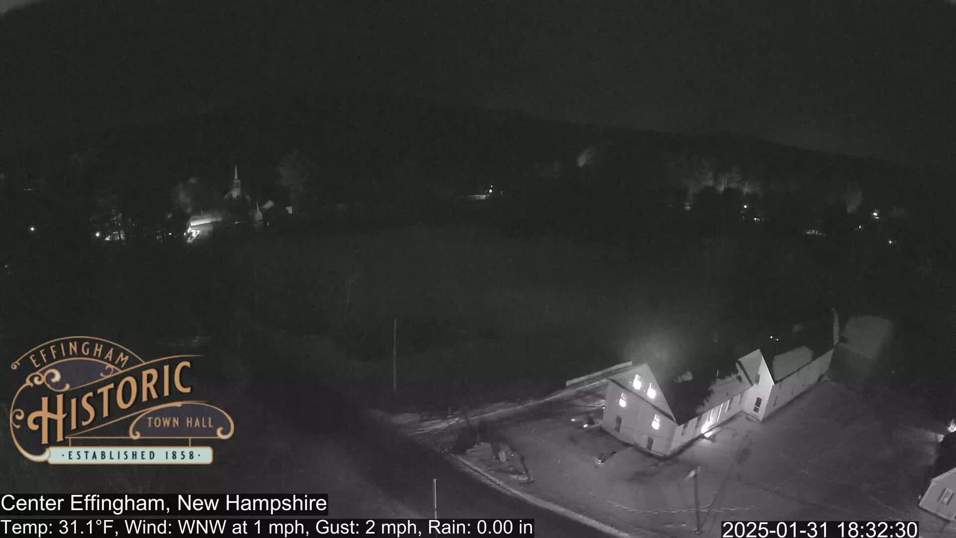 A nighttime, aerial view of a small town shows a building and a lightly snow-covered road under a dark, rainy sky.