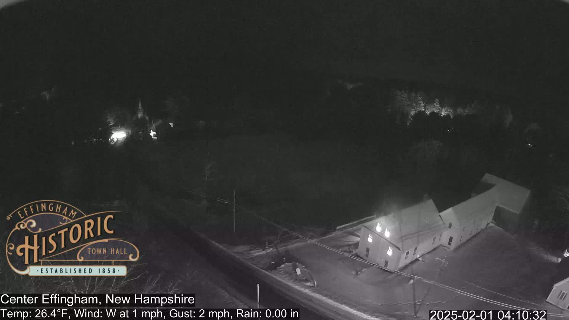 A nighttime, overhead view of a building complex and surrounding area with light snow cover and a west wind.