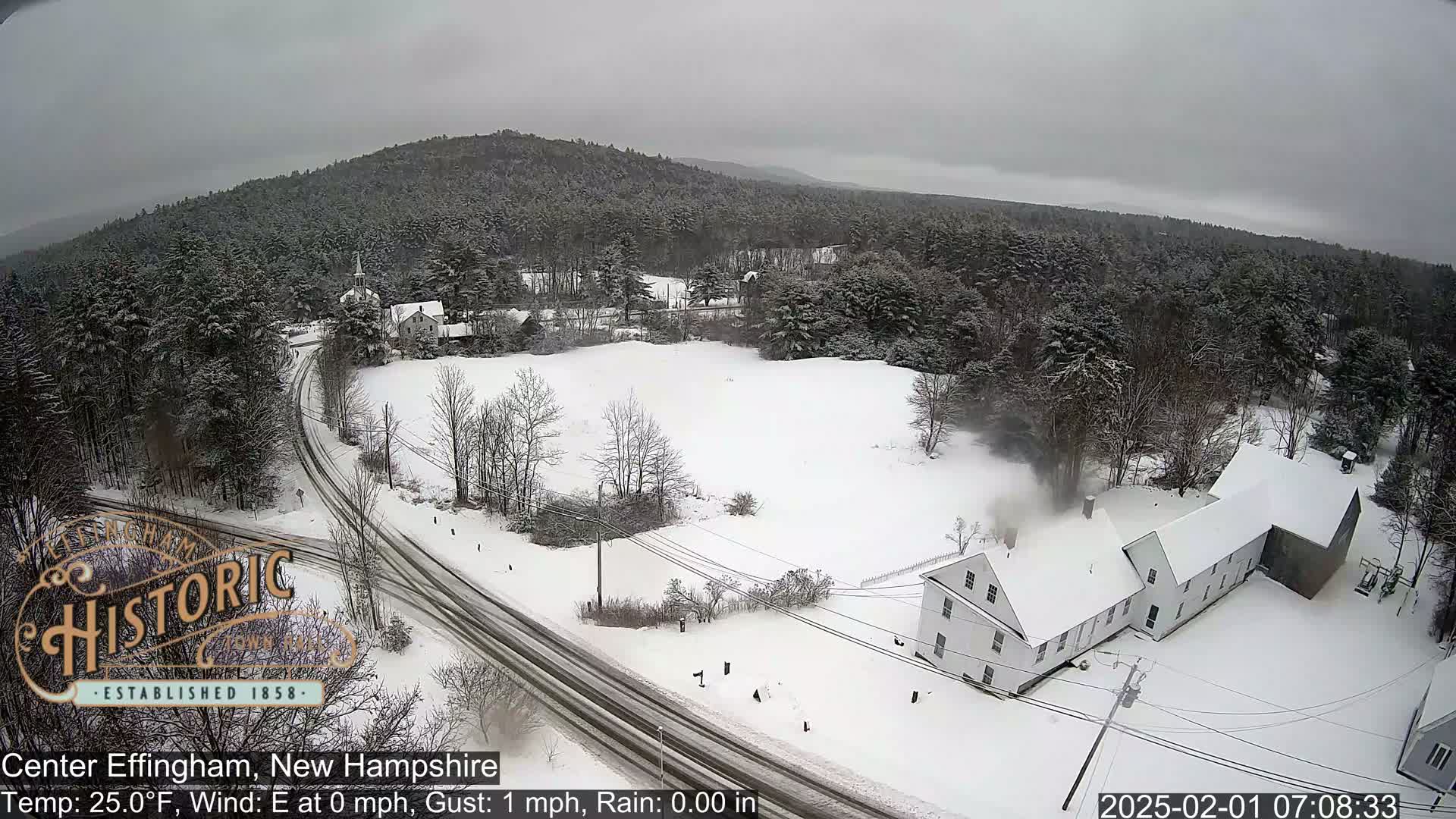 A snow-covered landscape shows a road curving through a field towards a long, white building, with a forested hillside in the background under an overcast sky.
