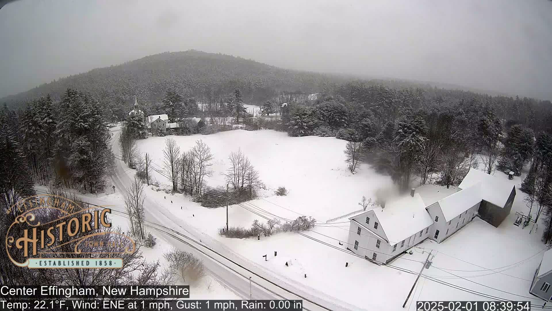 A snow-covered landscape with a white building and roads, under an overcast sky during a light snowfall.