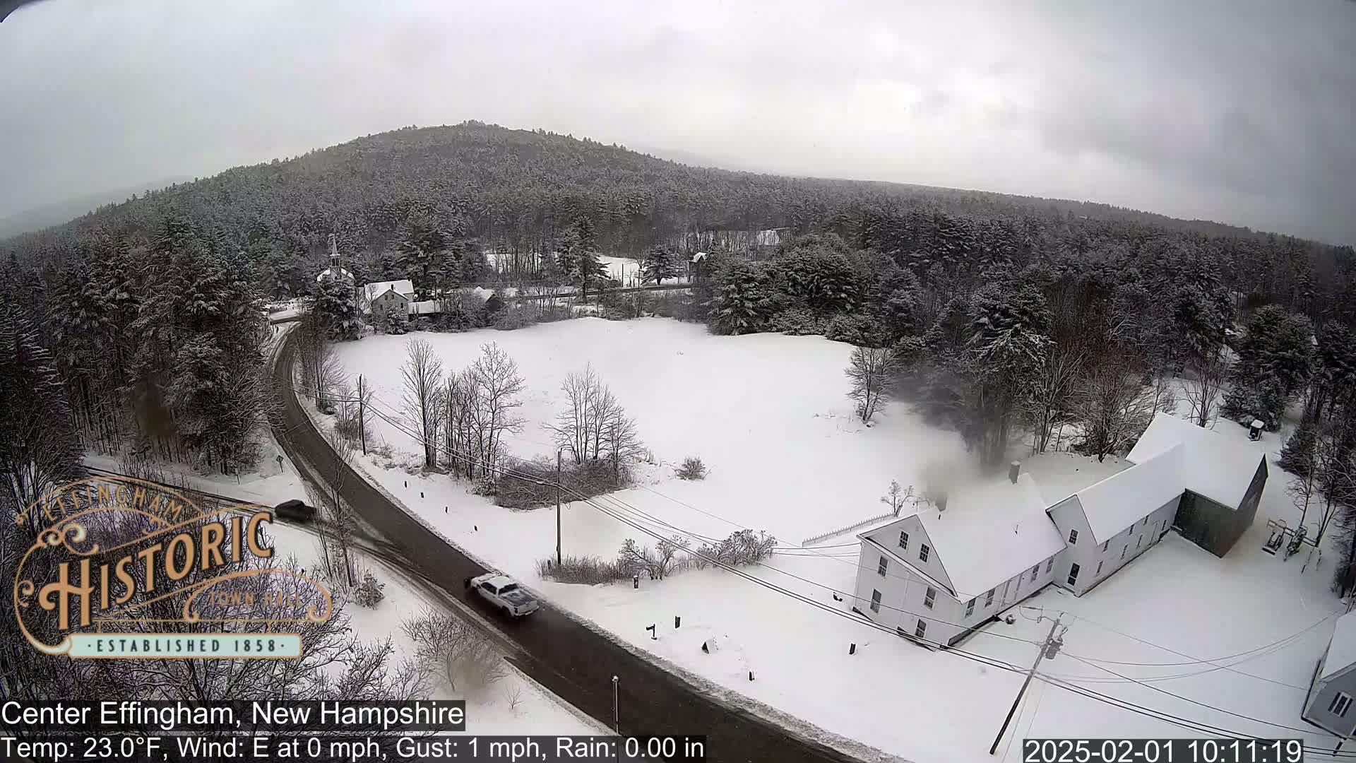 A snow-covered landscape with a road, a few buildings, and a wooded hill in the background, under light snow.