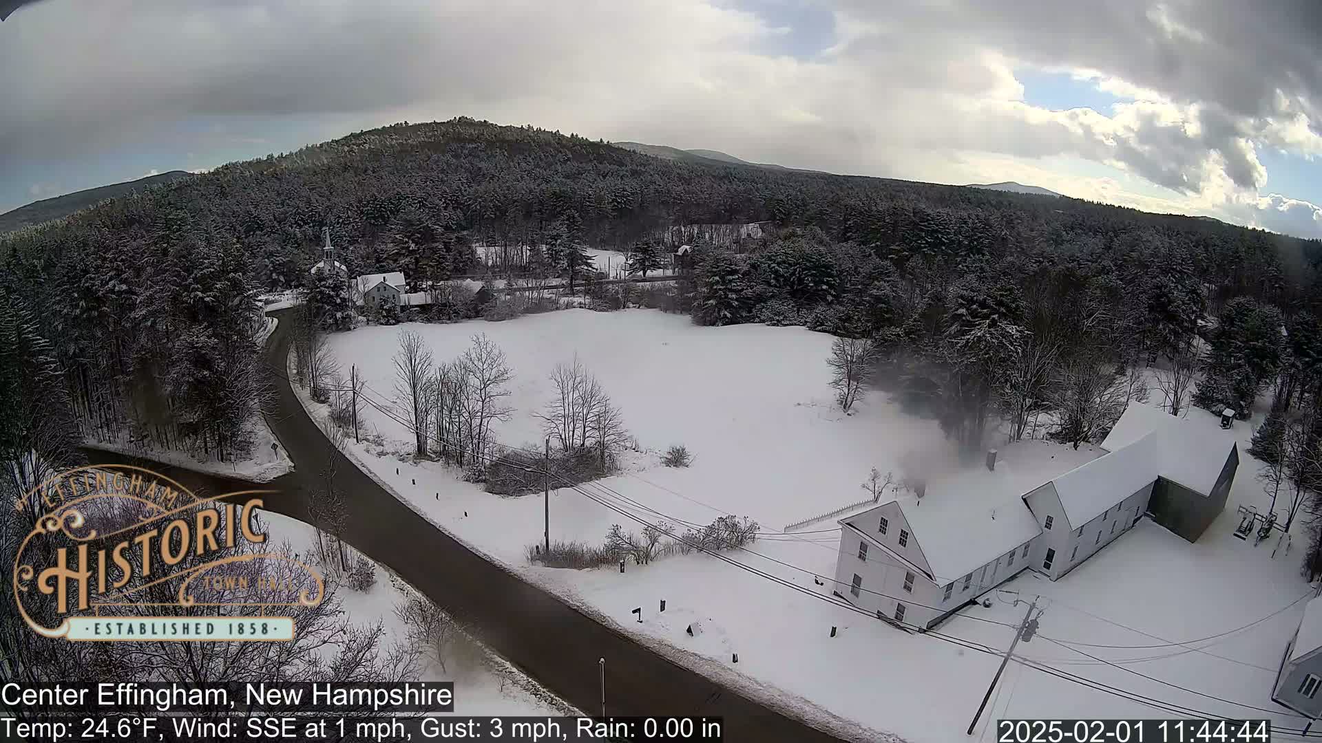 A snow-covered road winds through a winter landscape of snow-covered trees and buildings, under a partly cloudy sky.