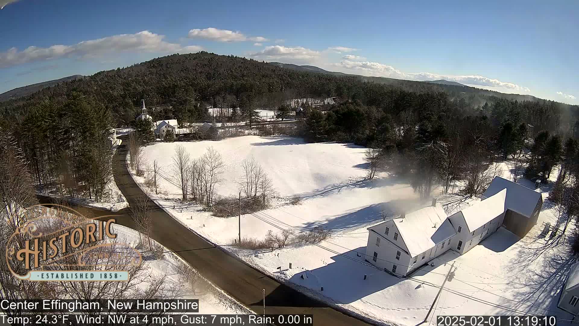 A snow-covered landscape with a road, buildings, and trees under a sunny sky.