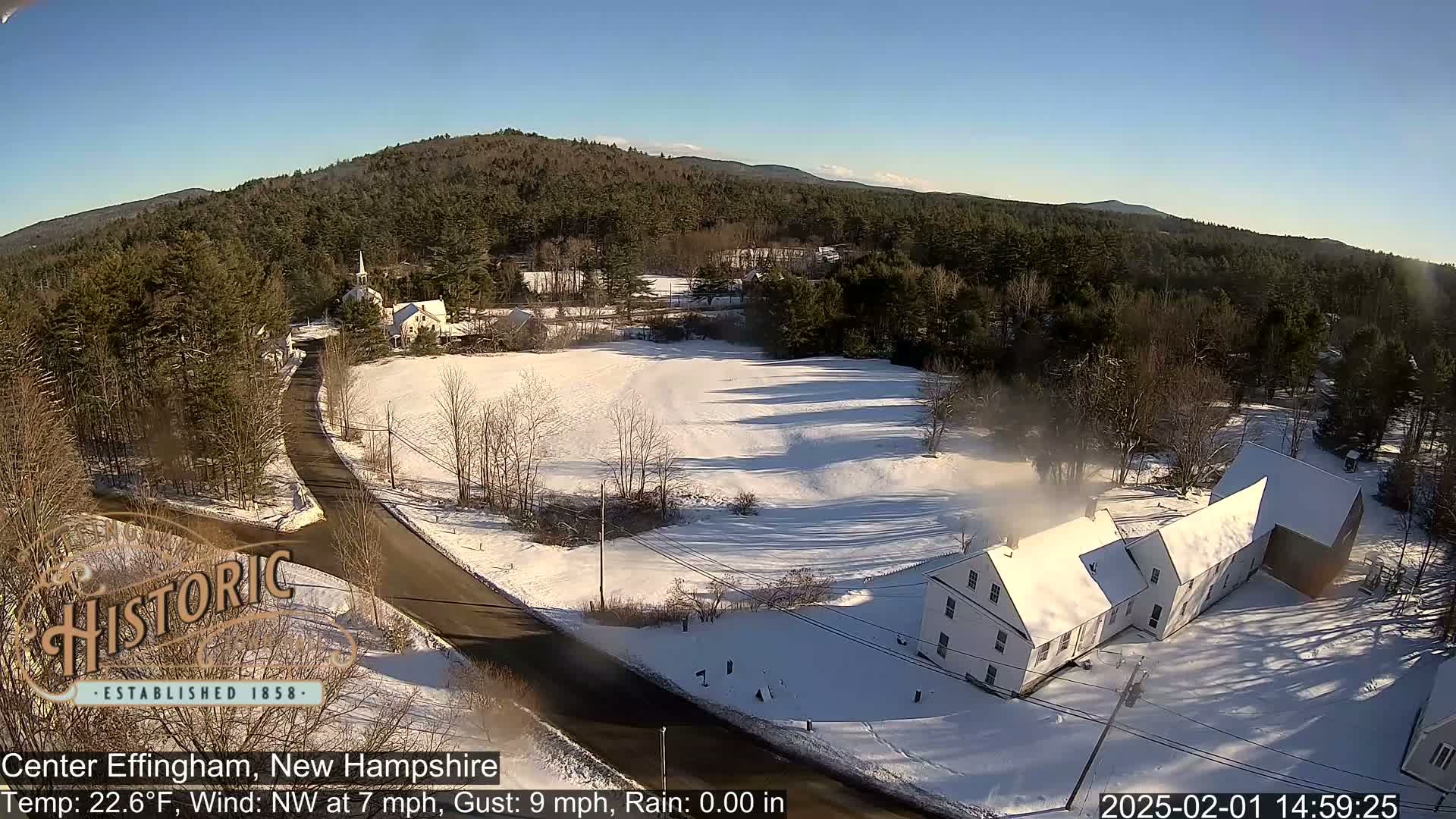 A snow-covered rural landscape with a road, buildings, and trees under a sunny sky.