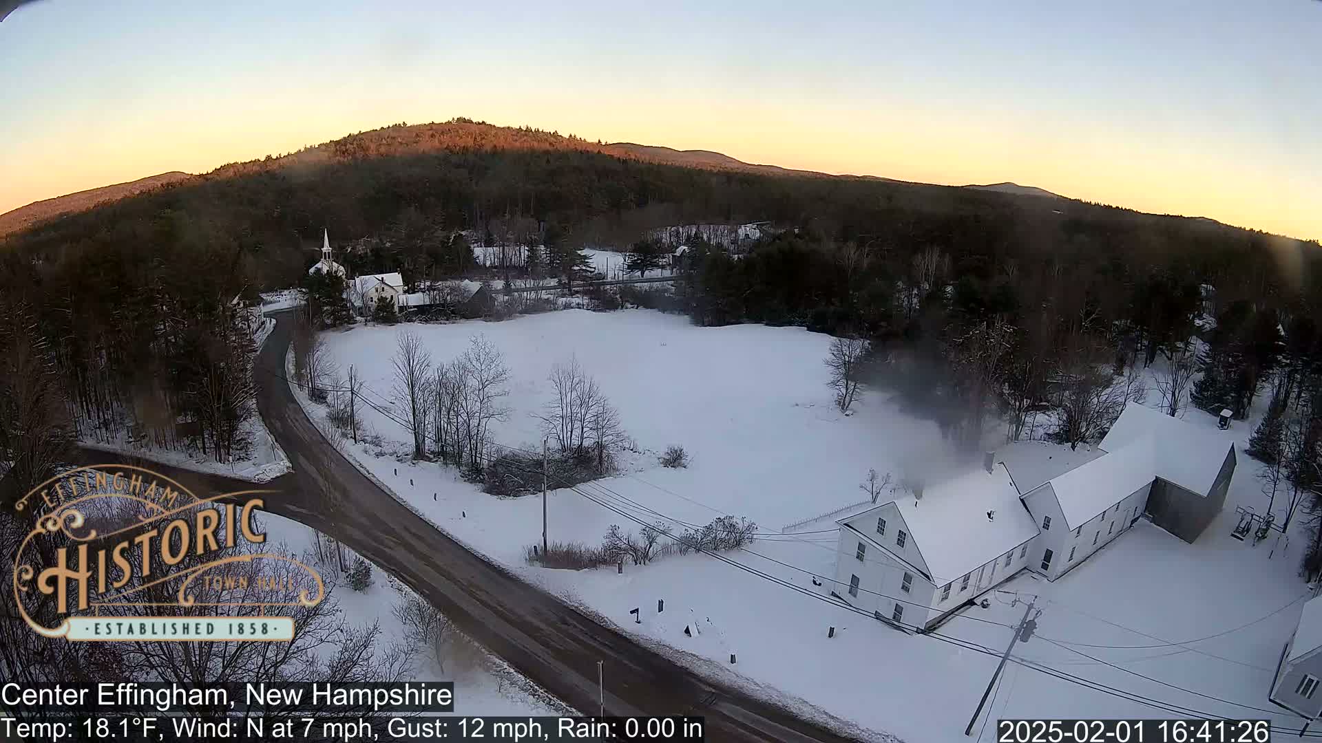 A snow-covered rural landscape at sunset, featuring a road, buildings, and a church, with light winds.