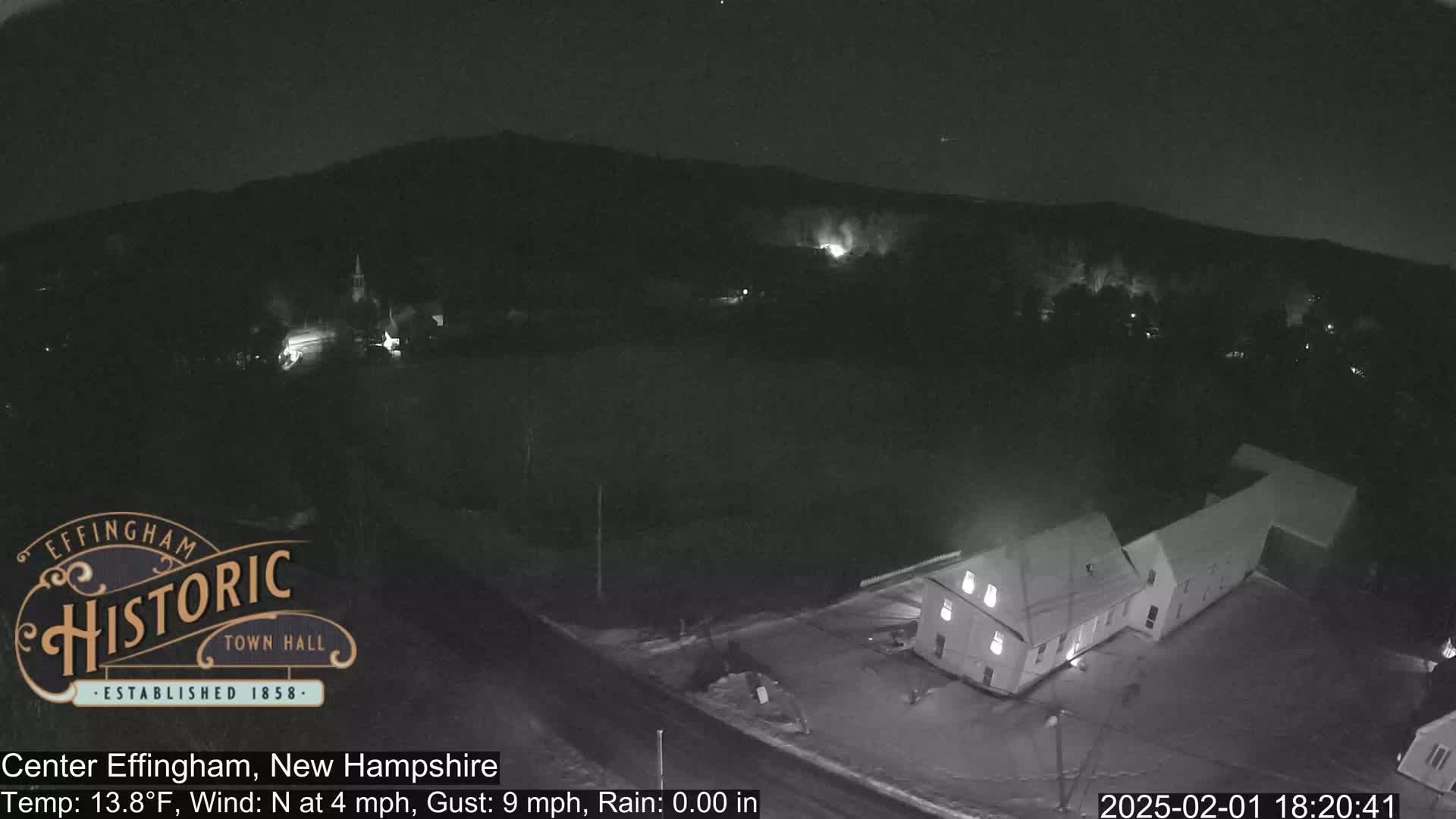 A nighttime, aerial view of a small town under a dark sky with a north wind, showing a building complex illuminated, and some sparsely lit areas in the distance.