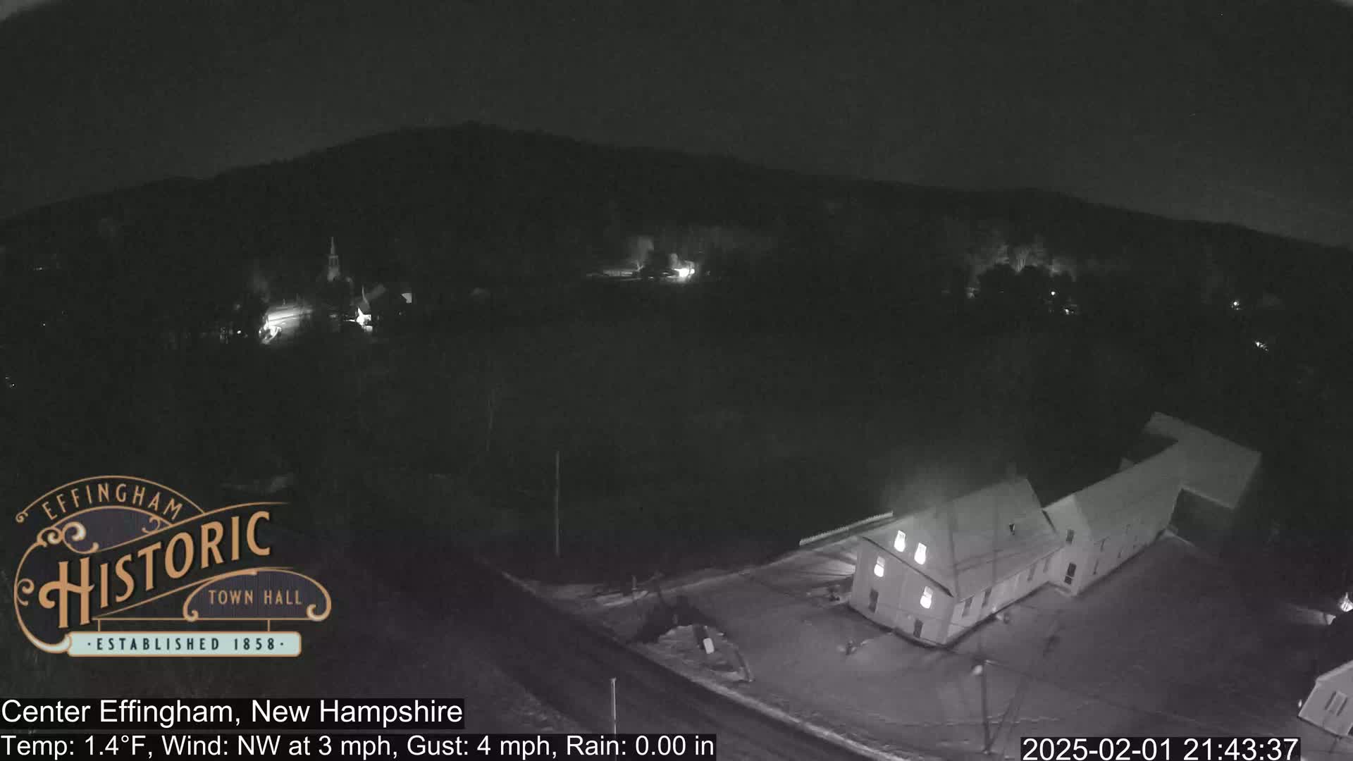 A nighttime, low-light aerial view of a small town with a few buildings, under a dark sky with a northwest wind and light rain.