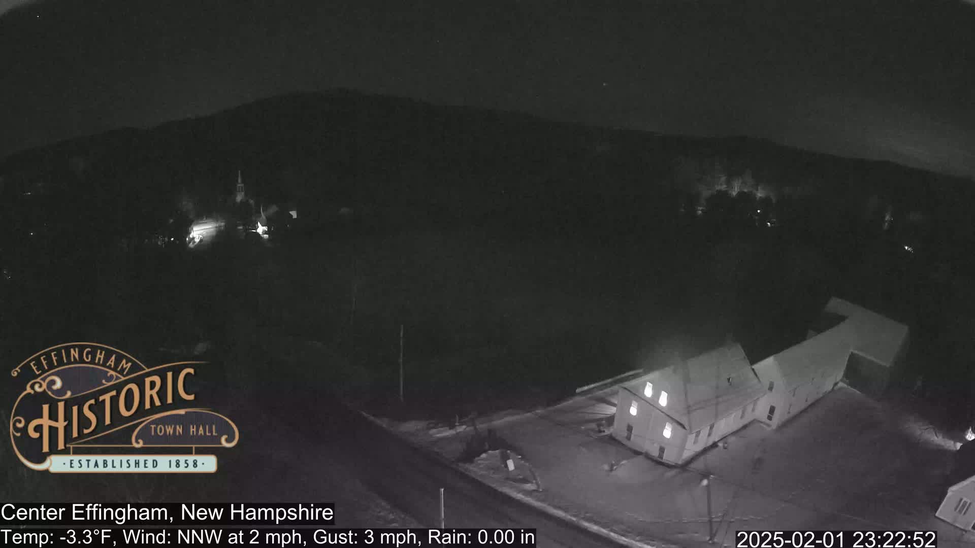 A nighttime, aerial view of a small town with a building lit up, under a clear, dark sky with a temperature of -3.3°F, a gentle north-northwest wind, and no rain.