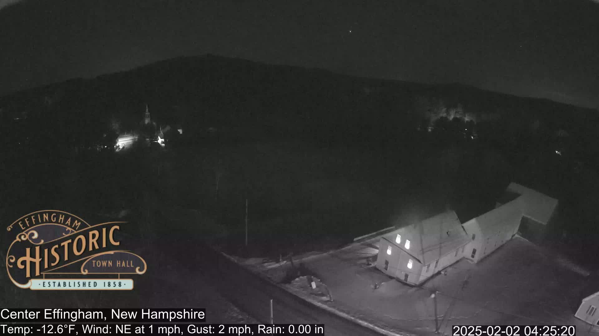 A nighttime aerial view of a small town shows a building illuminated against a dark, snowy landscape under a clear sky.