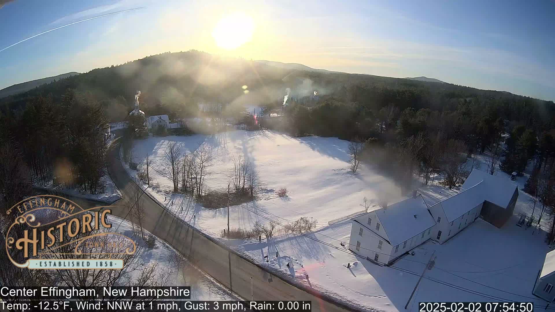 A snow-covered village scene is visible from an aerial perspective, under a sunny, cold morning sky.