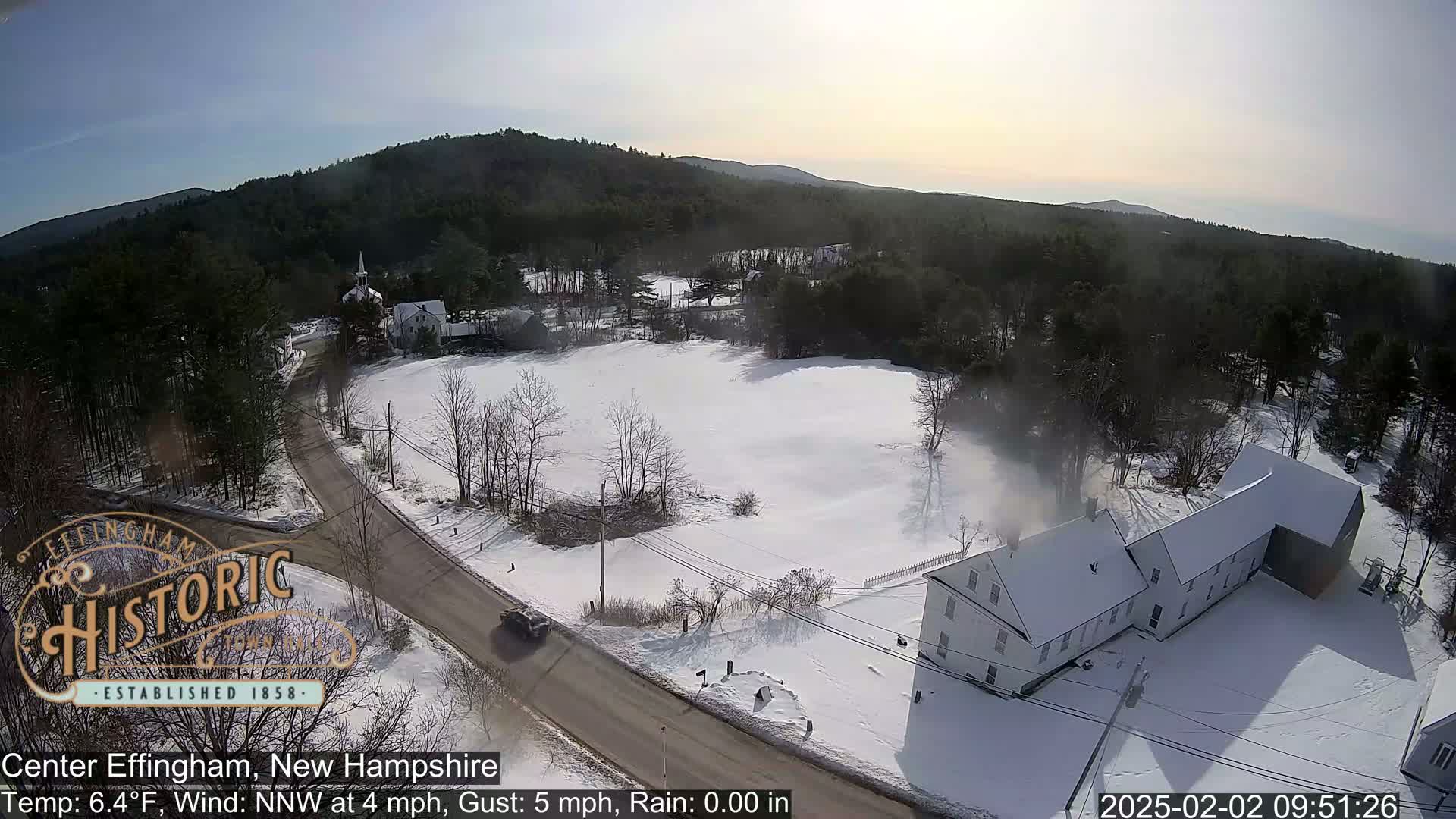 A snow-covered rural scene with a road, buildings, and trees under a partly sunny sky.