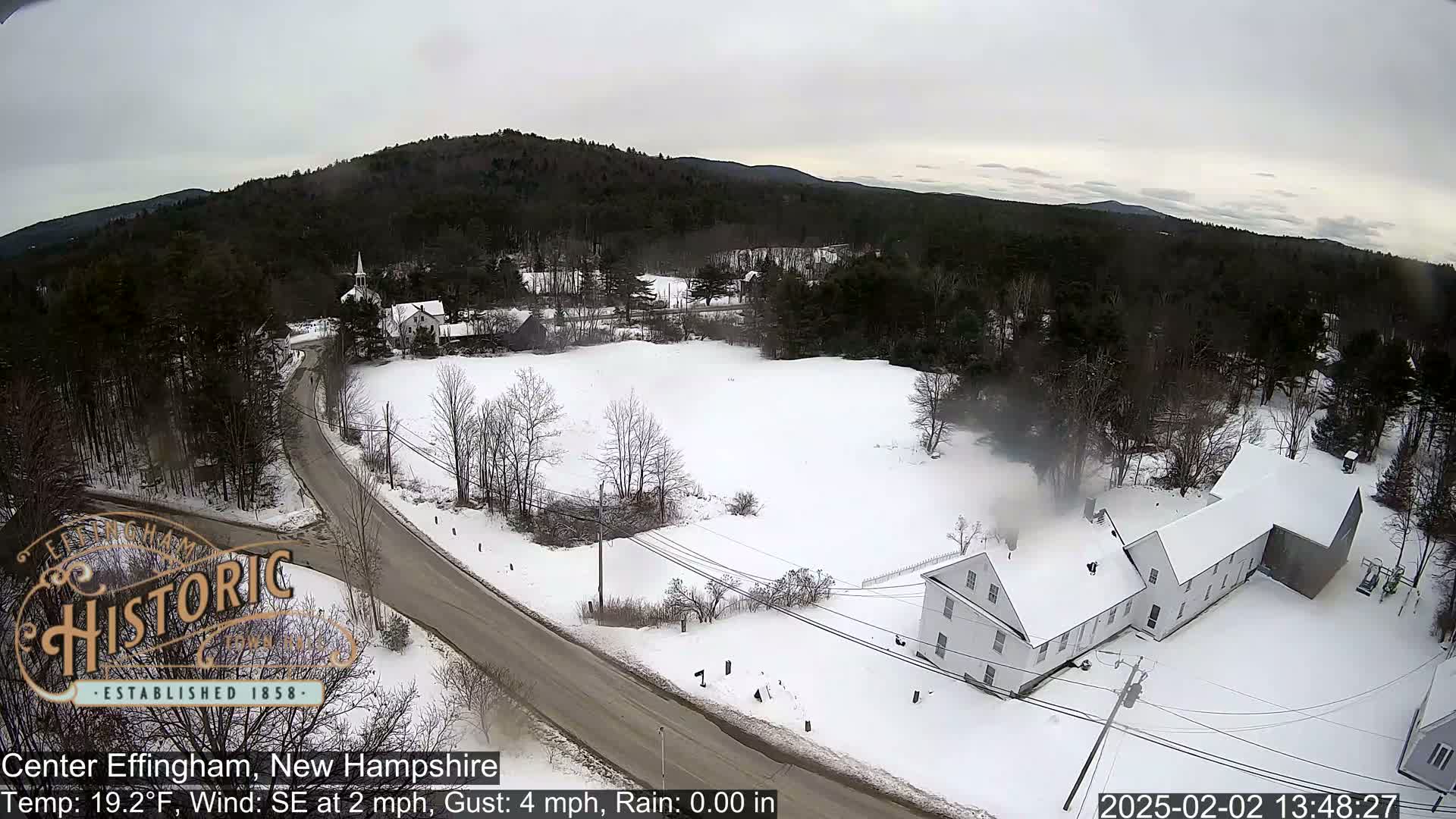 A snow-covered town with a road and buildings is visible from an aerial perspective under an overcast sky.