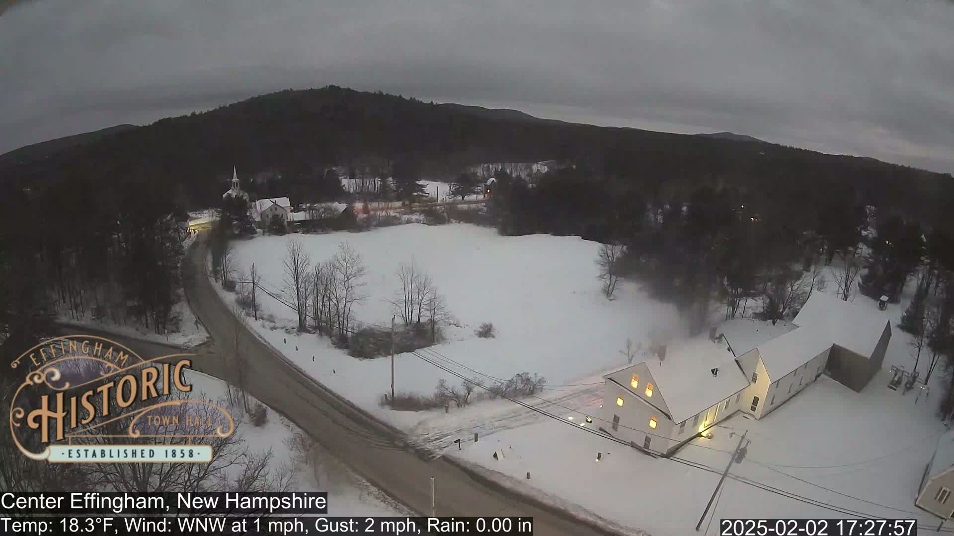 A snow-covered road winds through a small town nestled among dark, snow-dusted trees under a cloudy, overcast sky.