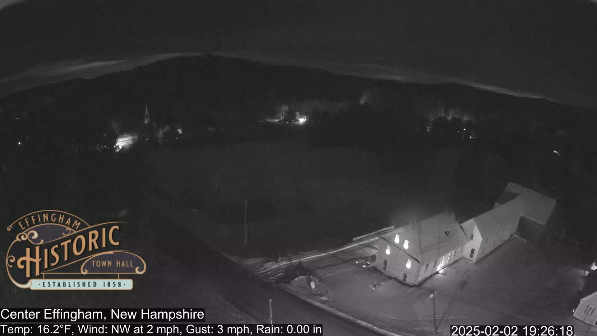 A nighttime aerial view of a snow-covered town with a building illuminated, under a dark sky with a northwest wind.
