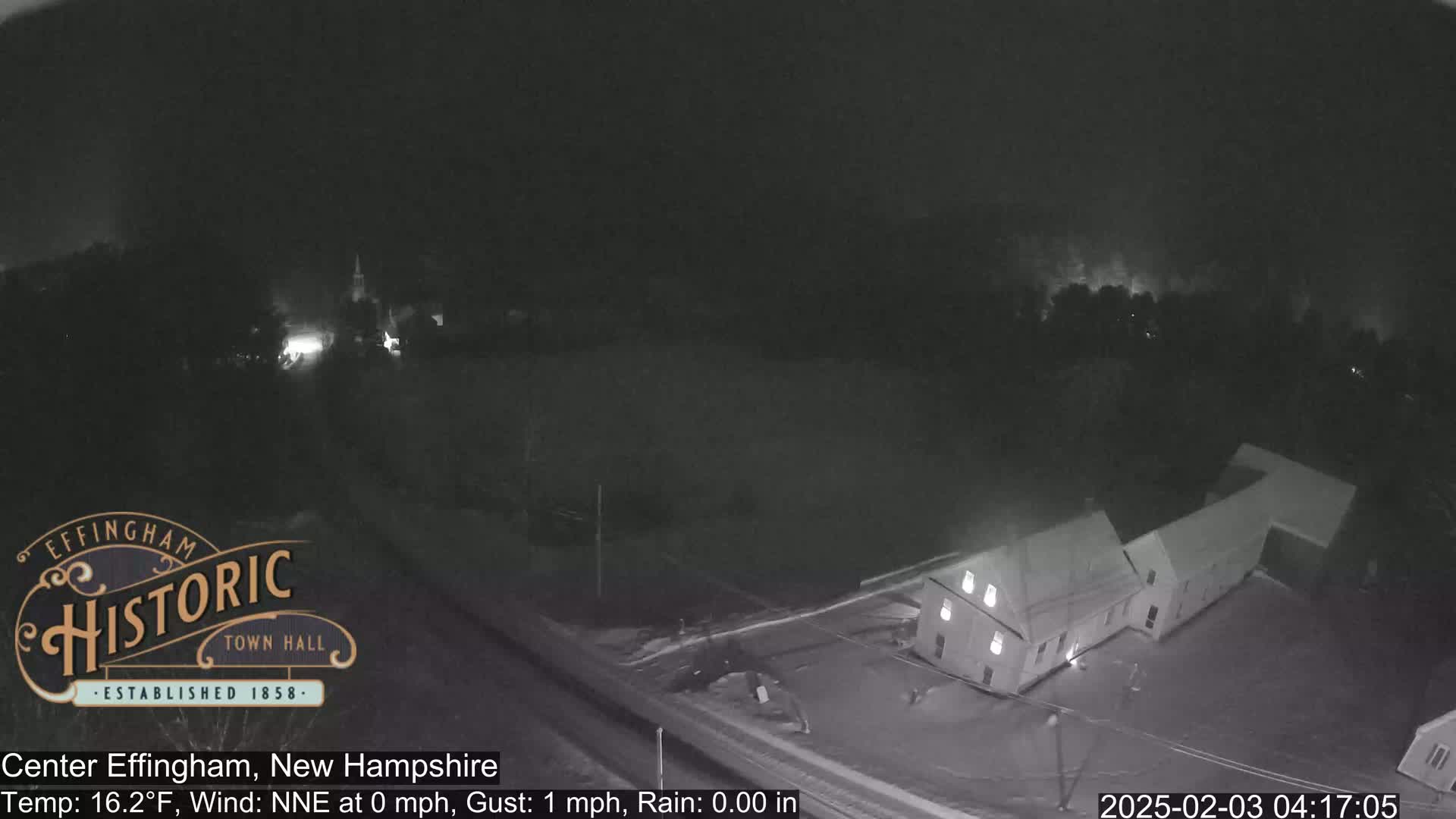 A nighttime, low-light view shows a building complex, possibly a town hall, with light snow or ice on the ground under a dark, overcast sky.