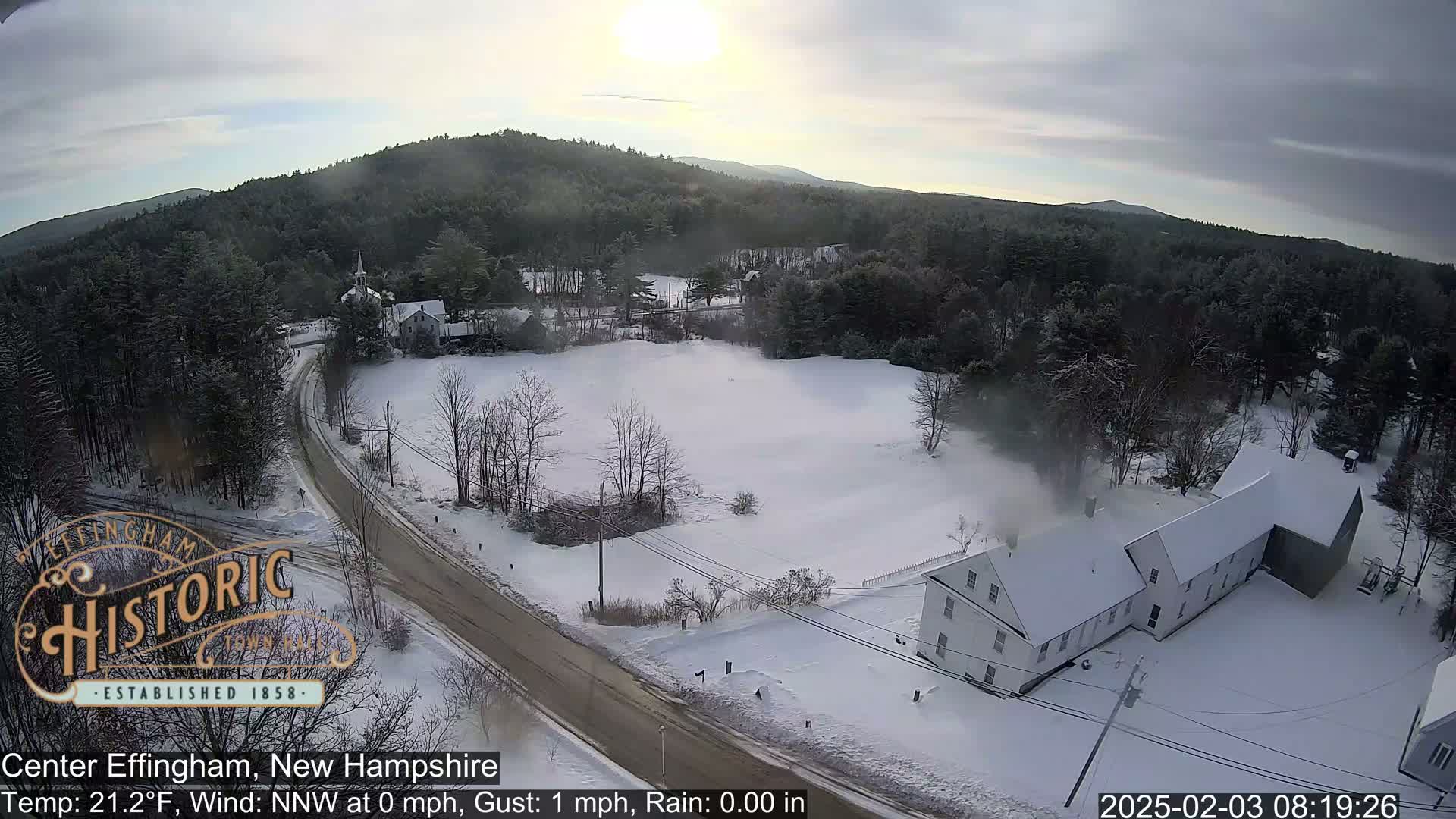 A snow-covered rural landscape with a road, buildings, and a partly sunny sky.