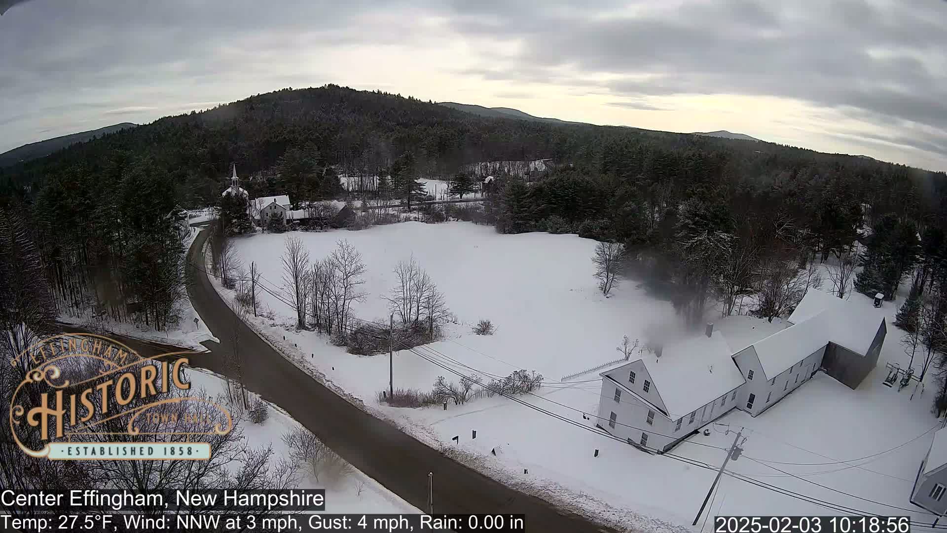 A snow-covered rural landscape with a road, buildings, and trees under a cloudy sky.