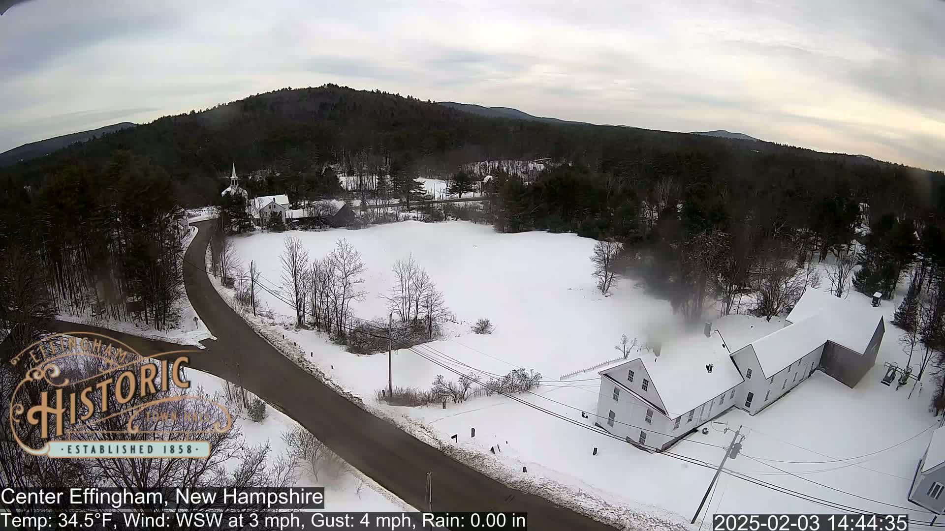 A snow-covered village with a road curving through it, nestled among bare trees and hills under an overcast sky.