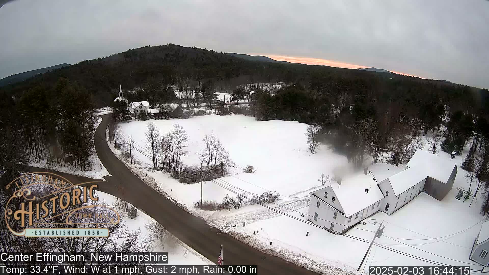 A snow-covered landscape shows a road intersecting with another, passing by a long white building and a church steeple, all under a cloudy, late-afternoon sky.