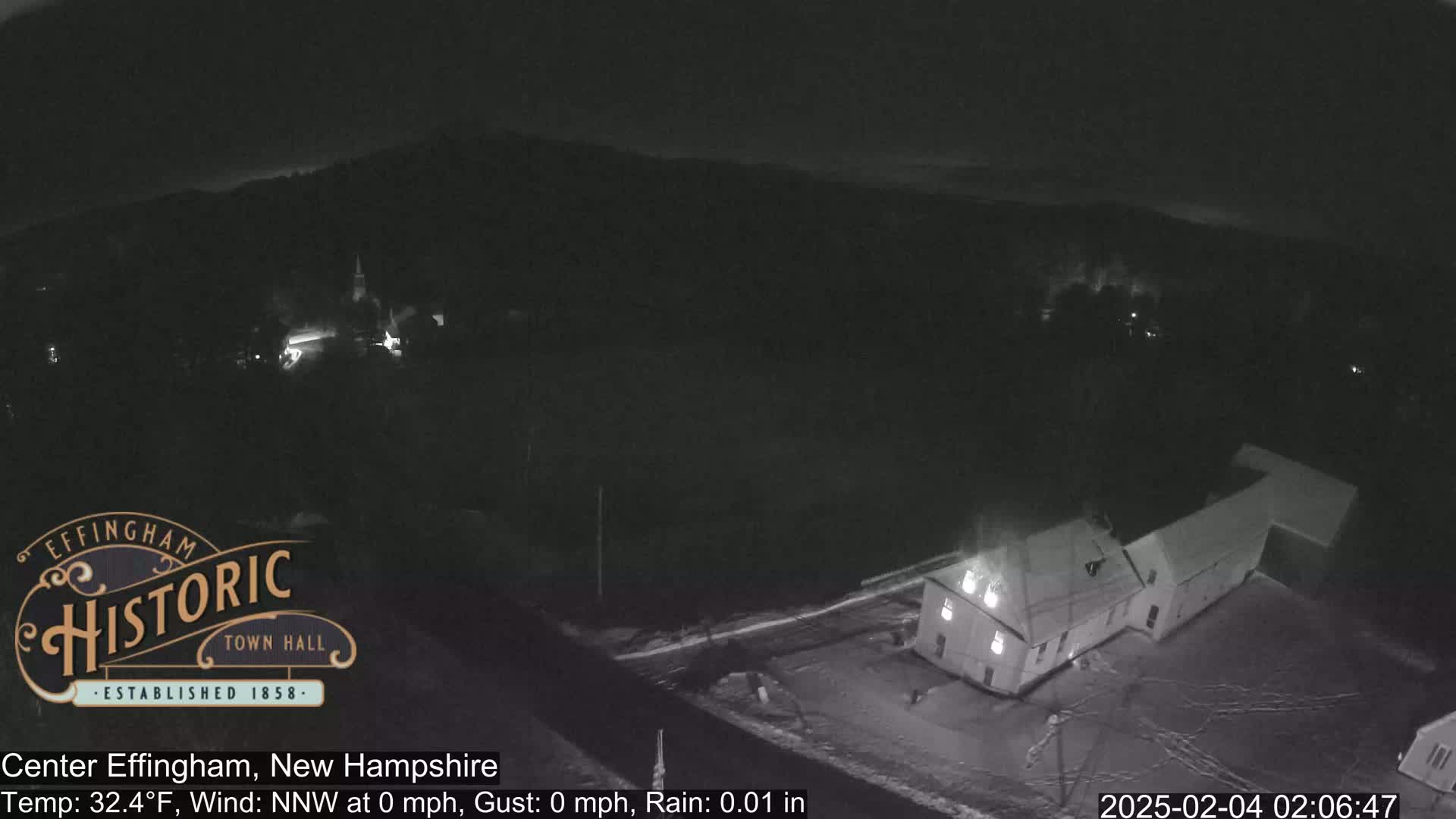 A nighttime, low-light aerial view of a snow-covered town, showing a building and a road under lightly raining conditions.