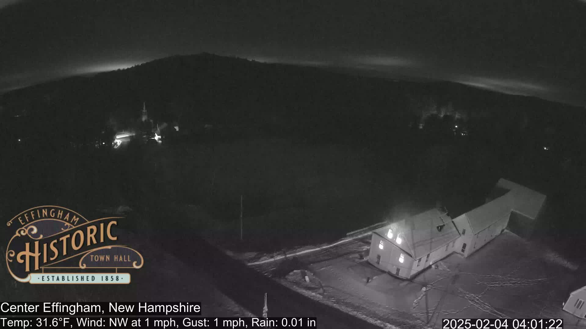 A nighttime, low-light aerial view of a snowy landscape showing a building and a road under a light rain with a slight northwest wind.
