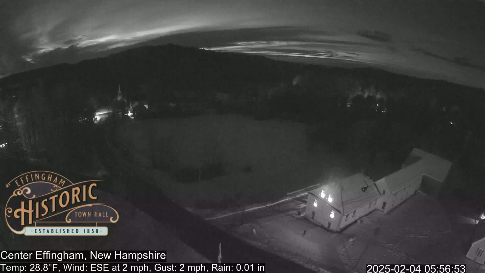 A nighttime aerial view of a building and a field under a partly cloudy sky with light rain.