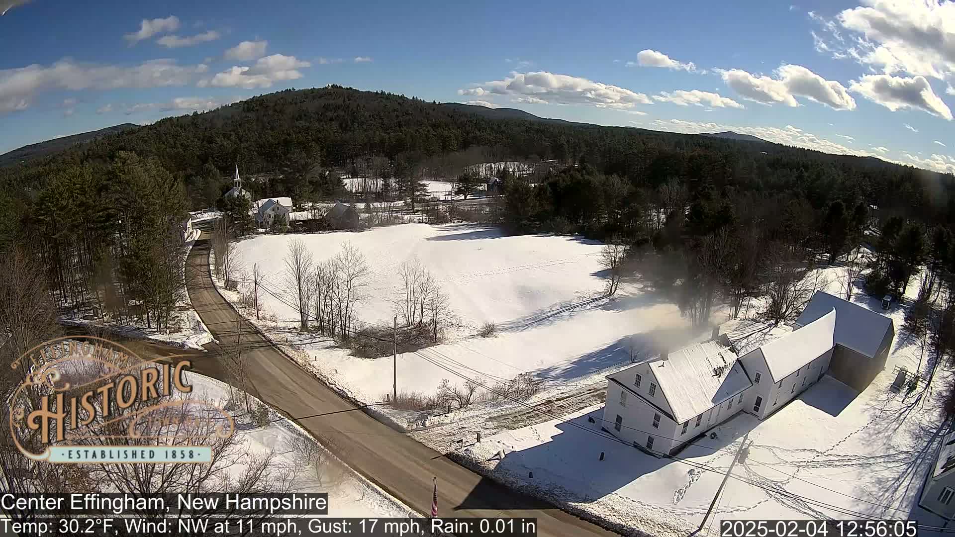 A snow-covered landscape with a road winding through a village, featuring a long, white building and forested hills under a partly cloudy sky.