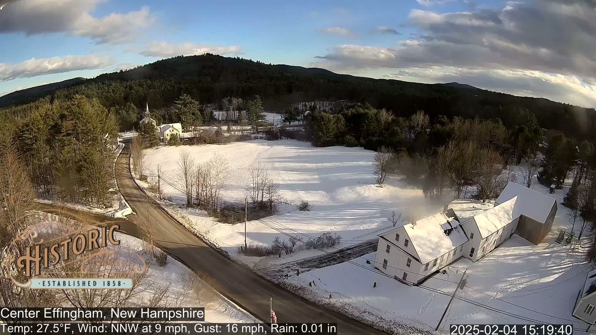 A snow-covered road winds through a small village nestled among snow-dusted trees and houses under a partly sunny sky.