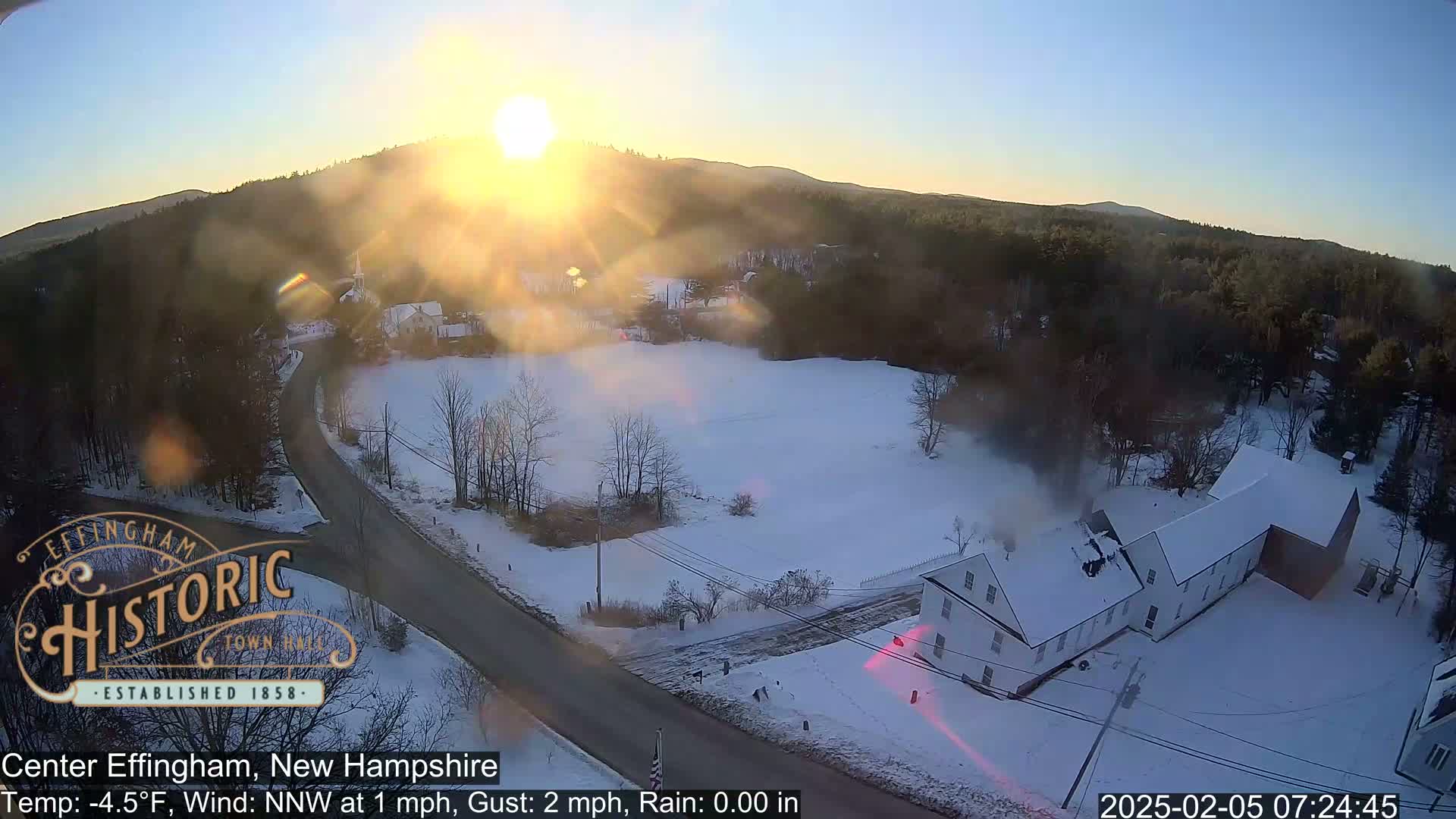 A snow-covered landscape at sunrise shows a road curving through a village with a long, white building, all under a mostly clear sky.